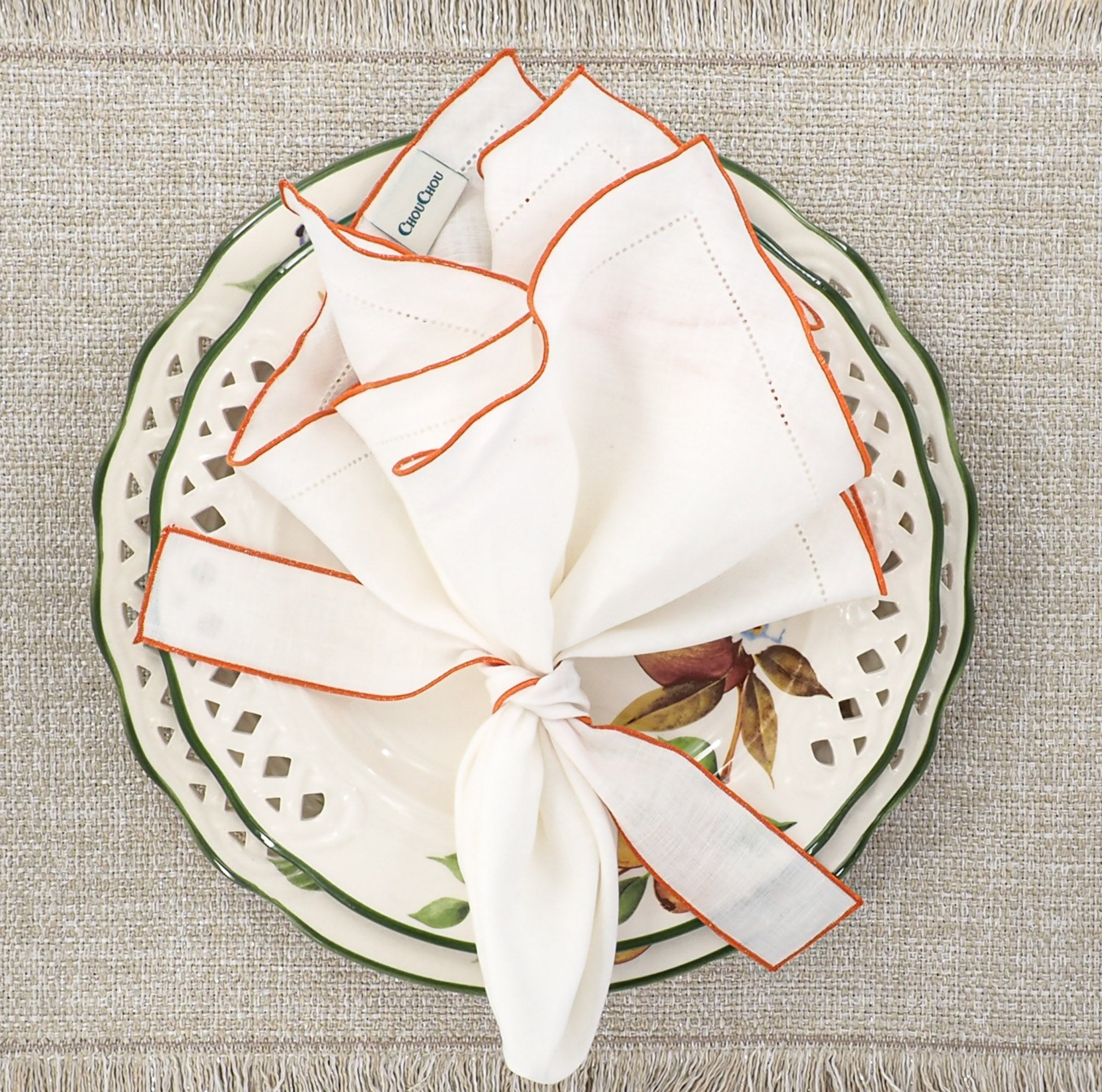 White linen napkins with orange ruffled hemstitched edge styled in a knot on a decorative plate, elegant table setting