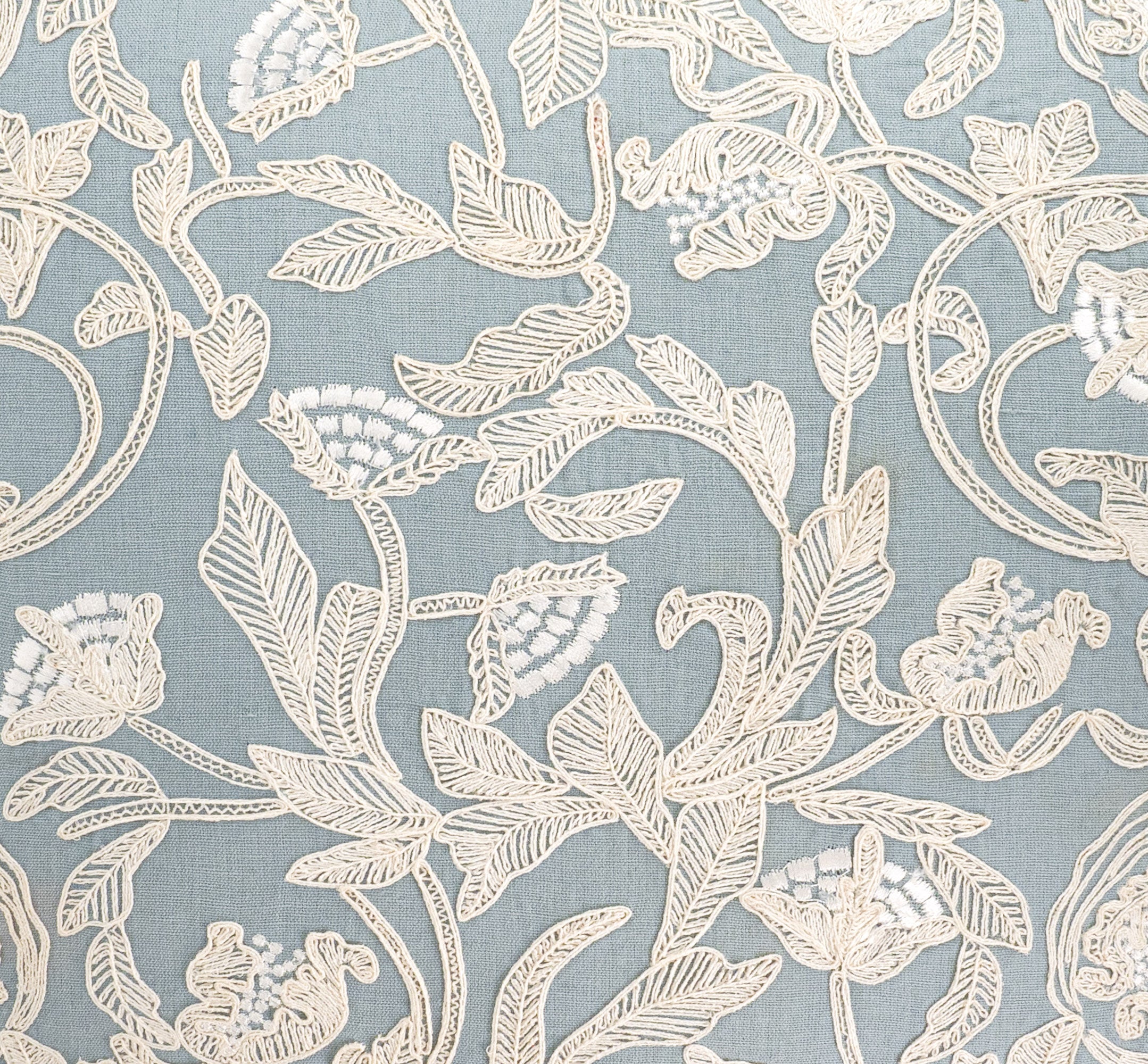 Close up of elegant square throw pillow with intricate ivory floral embroidery on soft blue linen.