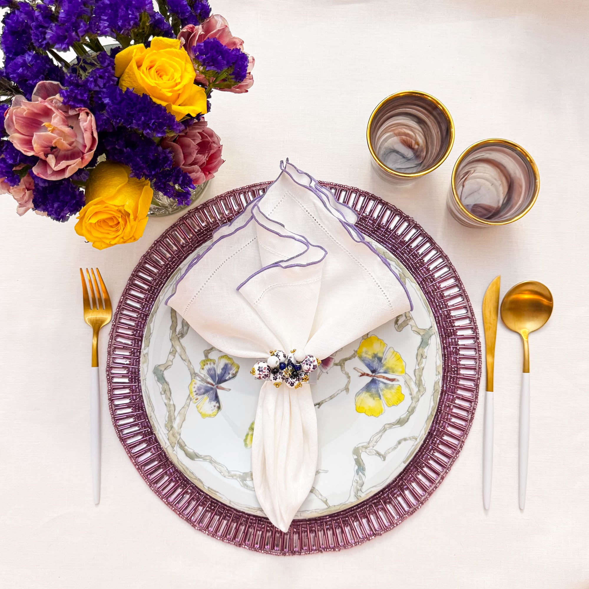 Handmade lavender beaded placemat styled with white linen napkin floral plate, and gold cutlery by ChouChou Touch