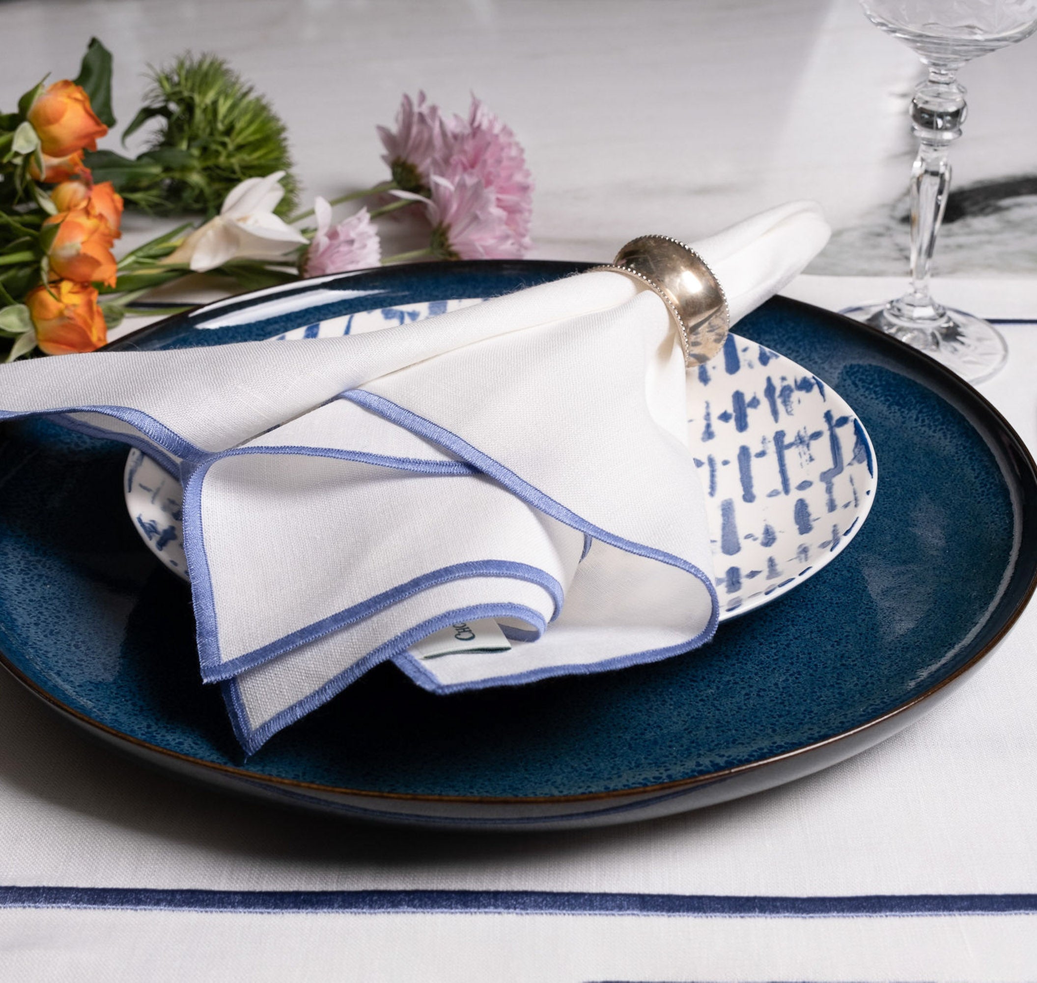 Elegant white linen napkins with blue stitched edges styled on layered blue dinnerware, creating a refined and timeless table setting with floral accents and soft luxury details.