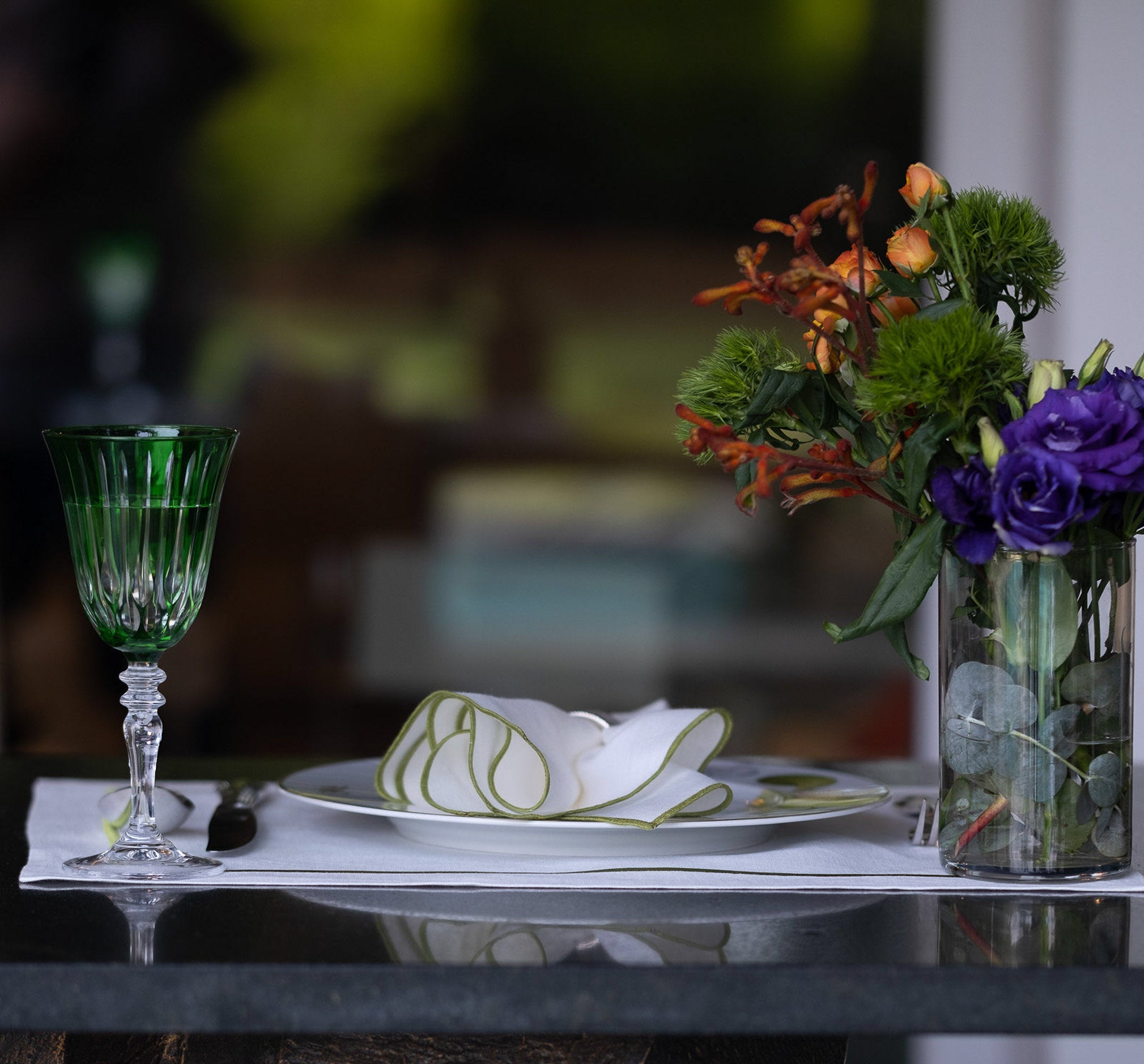 Close-up of elegant white linen napkins with delicate green stitched edges layered over textured blue dinnerware, highlighting refined craftsmanship and timeless table styling details.
