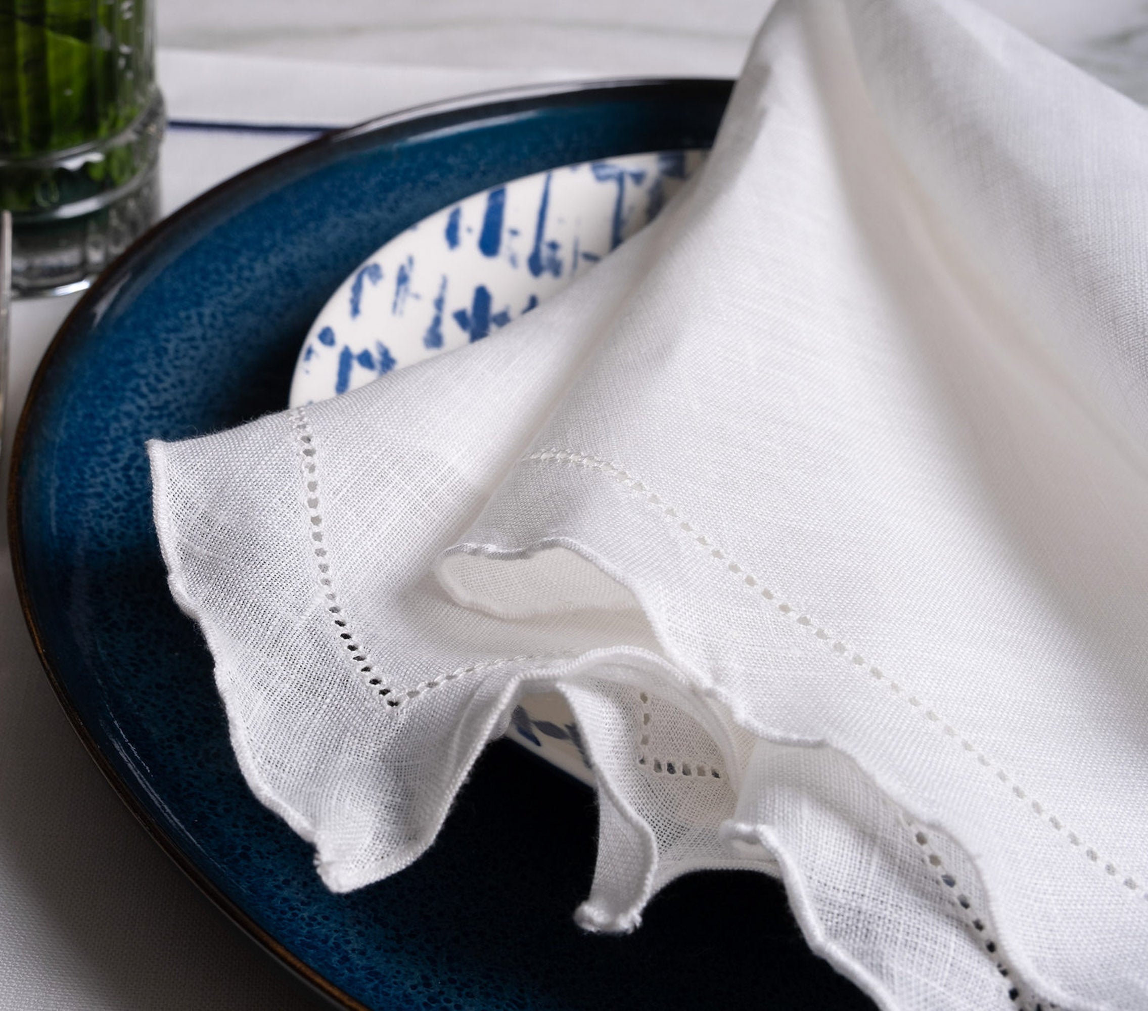 White linen napkin with ruffle hemstitched detail, layered with blue table linens for a sophisticated dining setting.