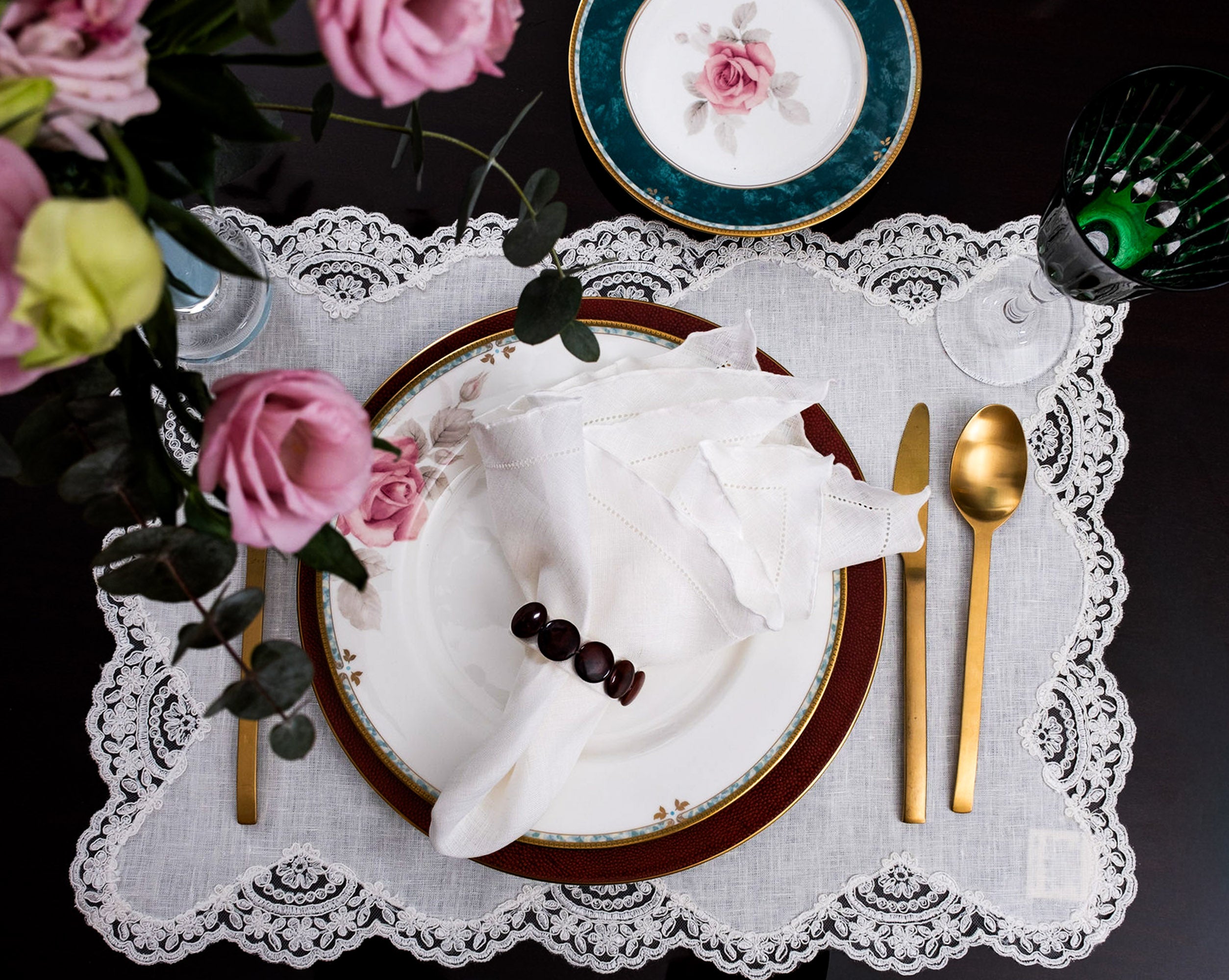 White linen ruffle hemstitched napkin styled on an elegant lace placemat with gold flatware and floral table setting on a dark background.
