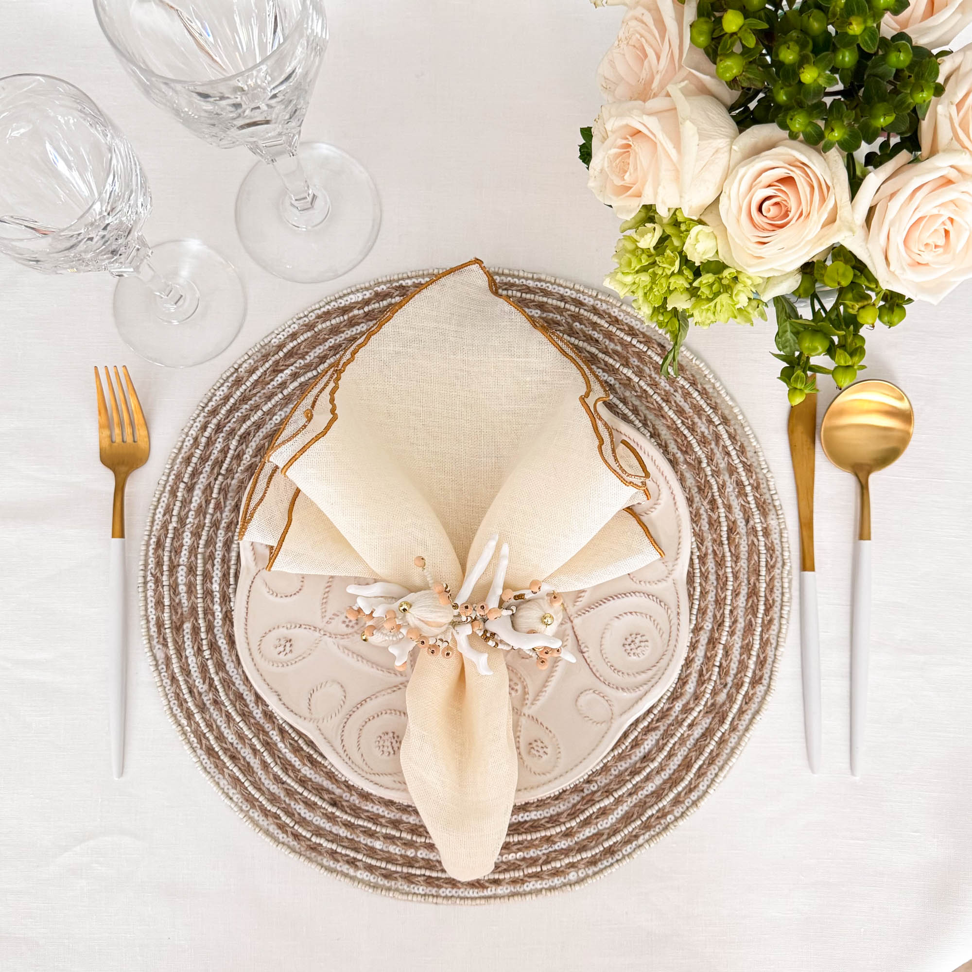 Elegant table setting featuring handmade braided jute placemat with white glass beads, styled ivory linen napkin with gold edges, floral napkin ring, gold cutlery, and white rose centerpiece by ChouChou Touch