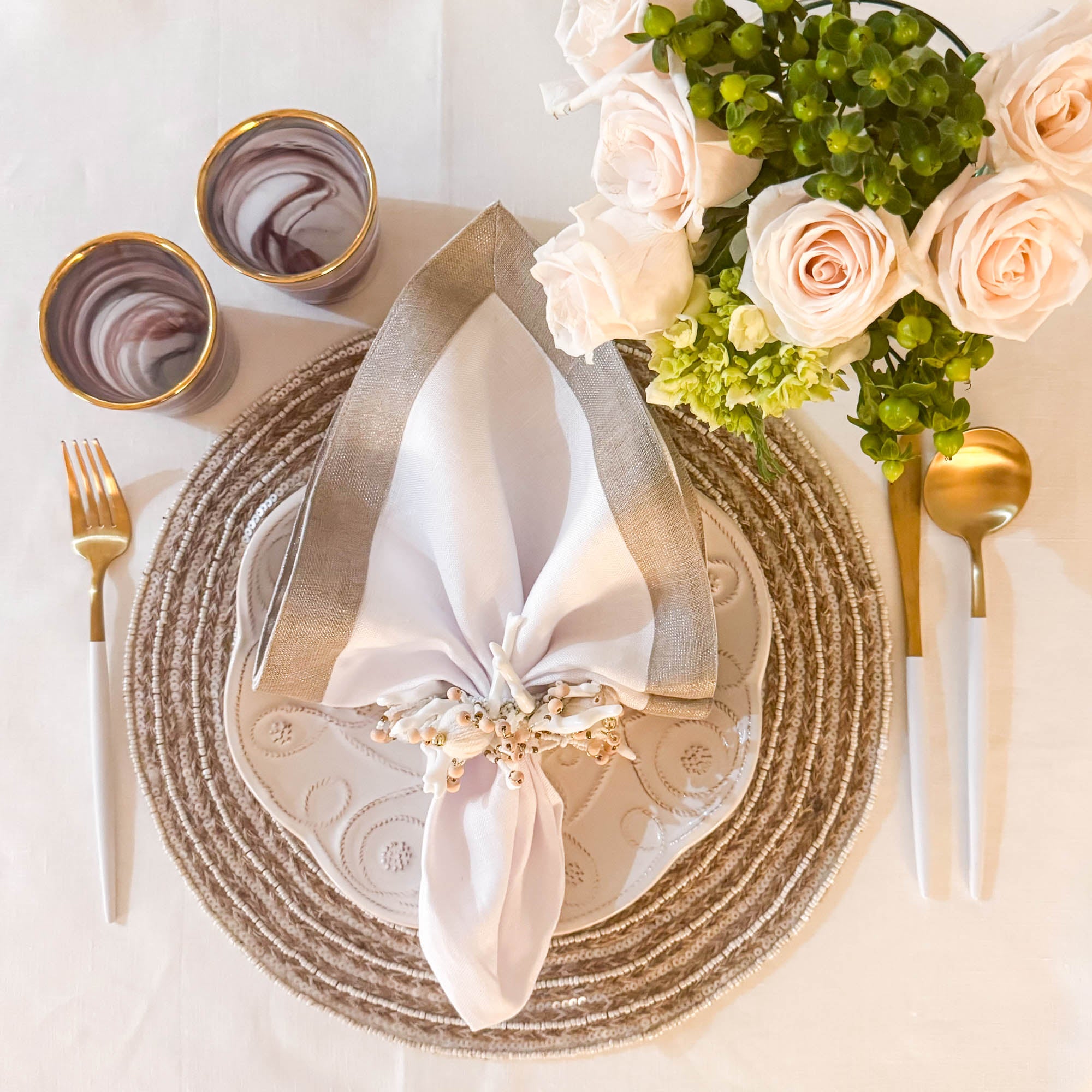 Sophisticated table decor featuring handmade jute placemat with white beadwork, paired with linen napkin, floral napkin ring, and gold cutlery by ChouChou Touch