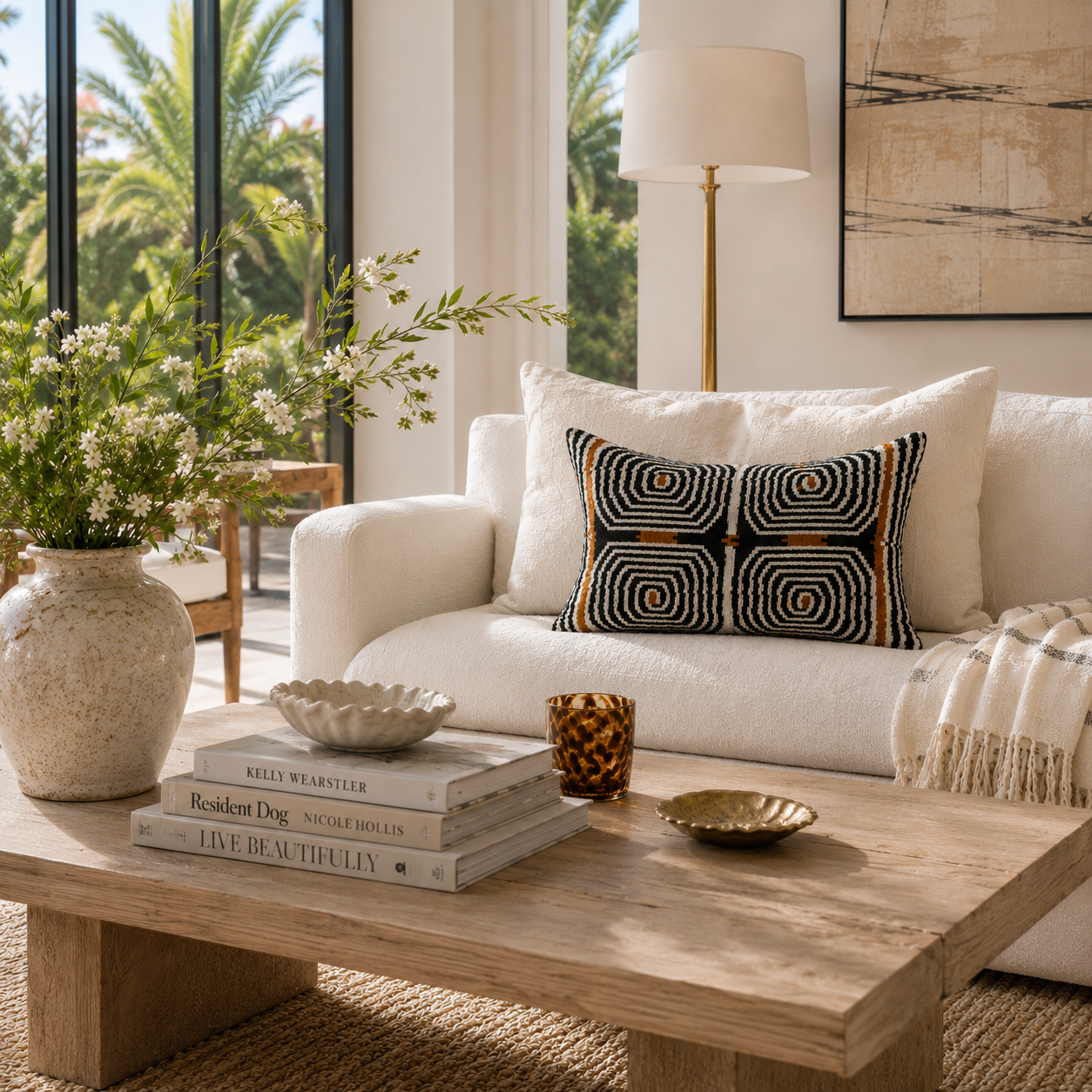 Handwoven silk velvet ikat lumbar pillow in black, ivory and bronze styled on an ivory sofa in a bright Palm Beach-inspired living room with natural wood, soft neutral decor and warm sunlight.
