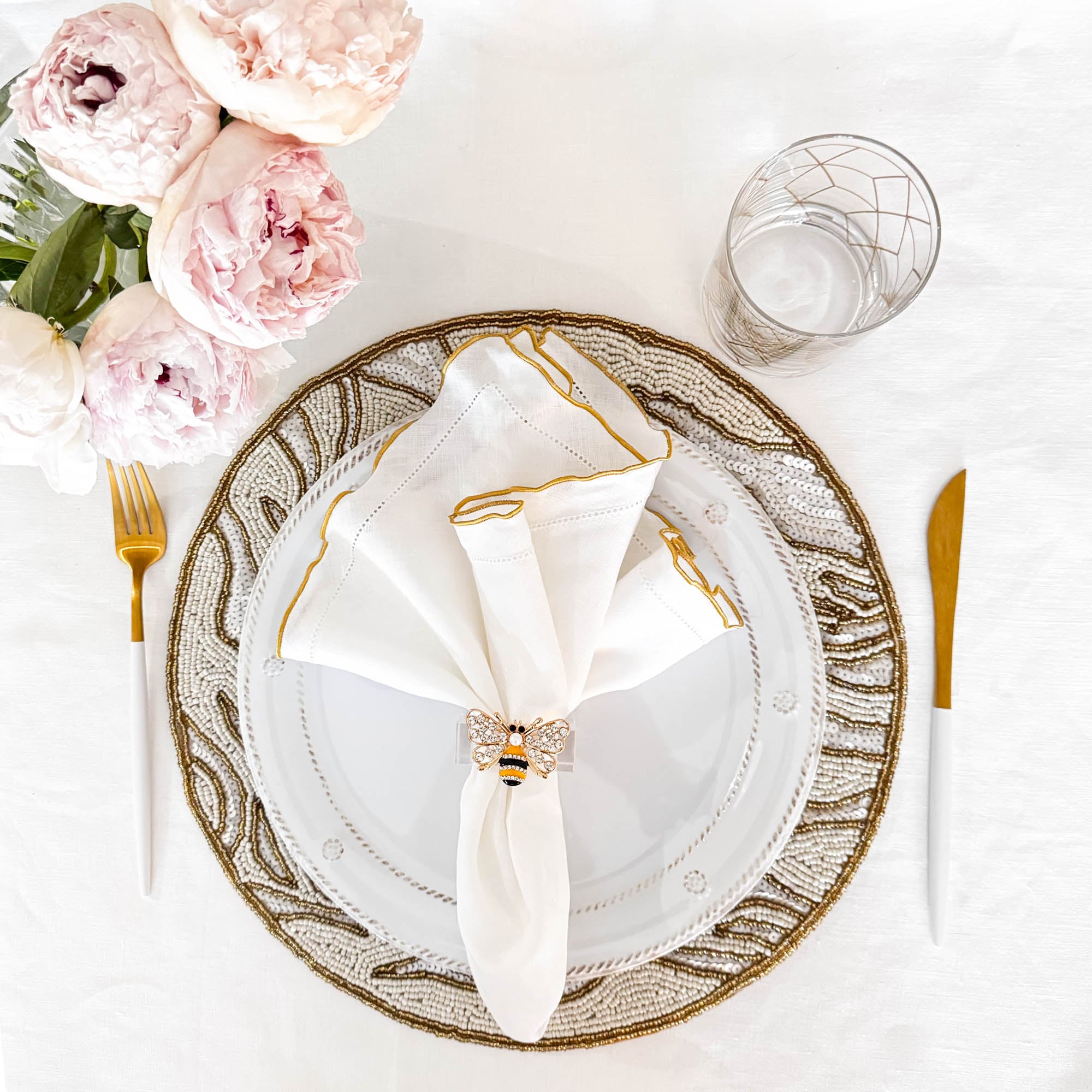 Decorative table setting with a white linen napkin with gold edged  and with bee  napkin ring, and black and gold brooch on a white blinen tablecloth background.