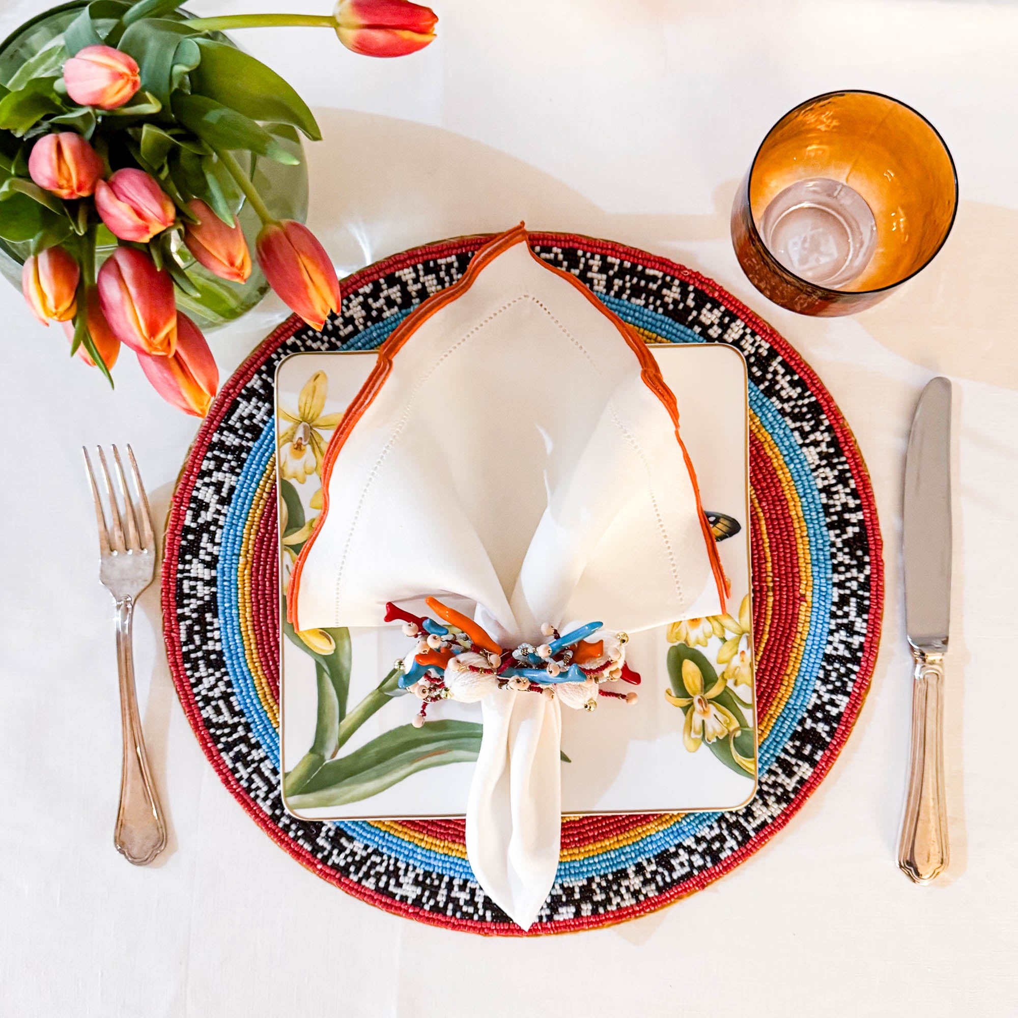 white linen napkin with orange ruffle hemstitched edge styled on a colorful decorative plate with beaded napkin ring, tulips and glassware, elegant table setting