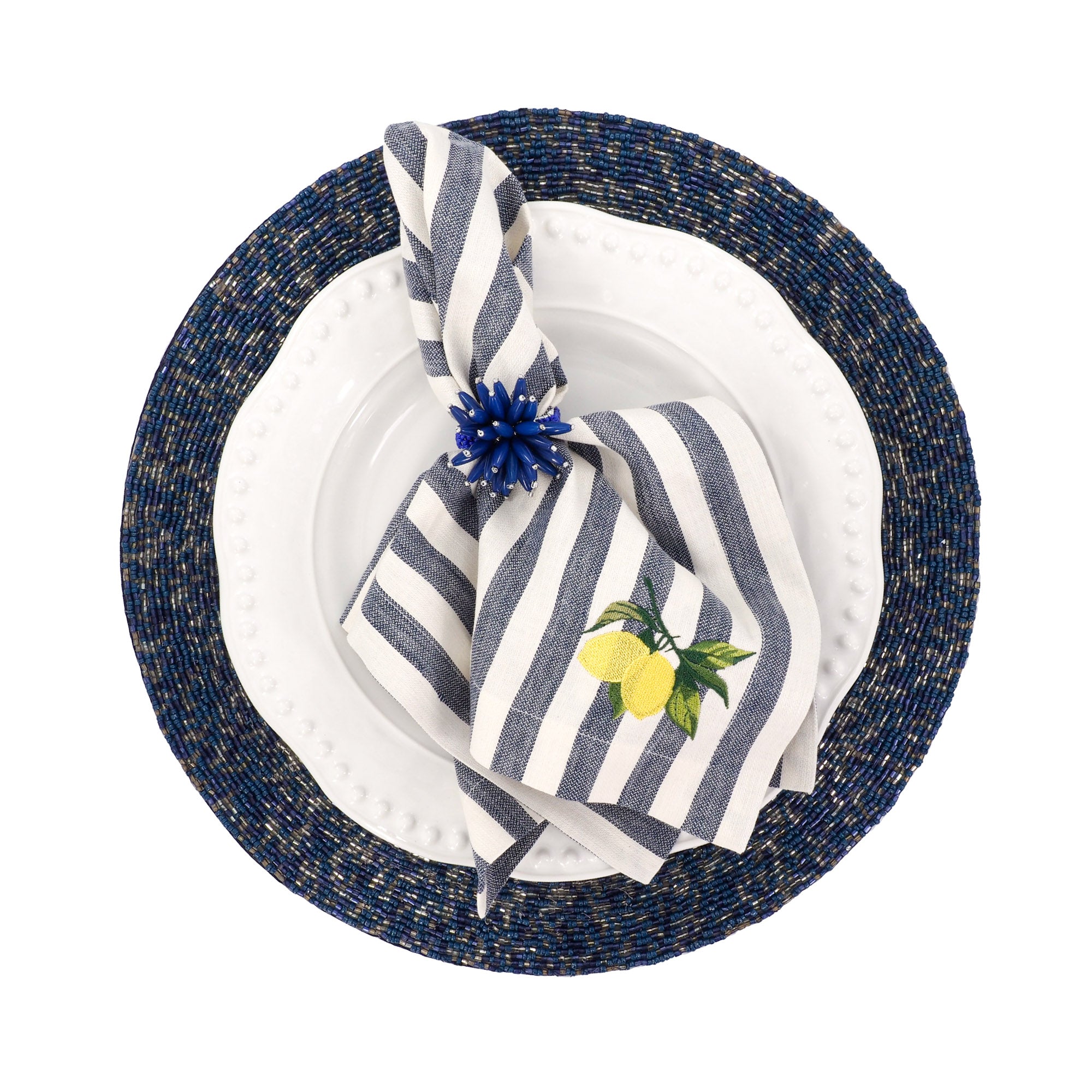 A top-down view of a place setting featuring a round navy blue woven placemat, a white plate, a folded navy and white striped napkin with a yellow lemon illustration, and a blue  napkin holder on a white background.