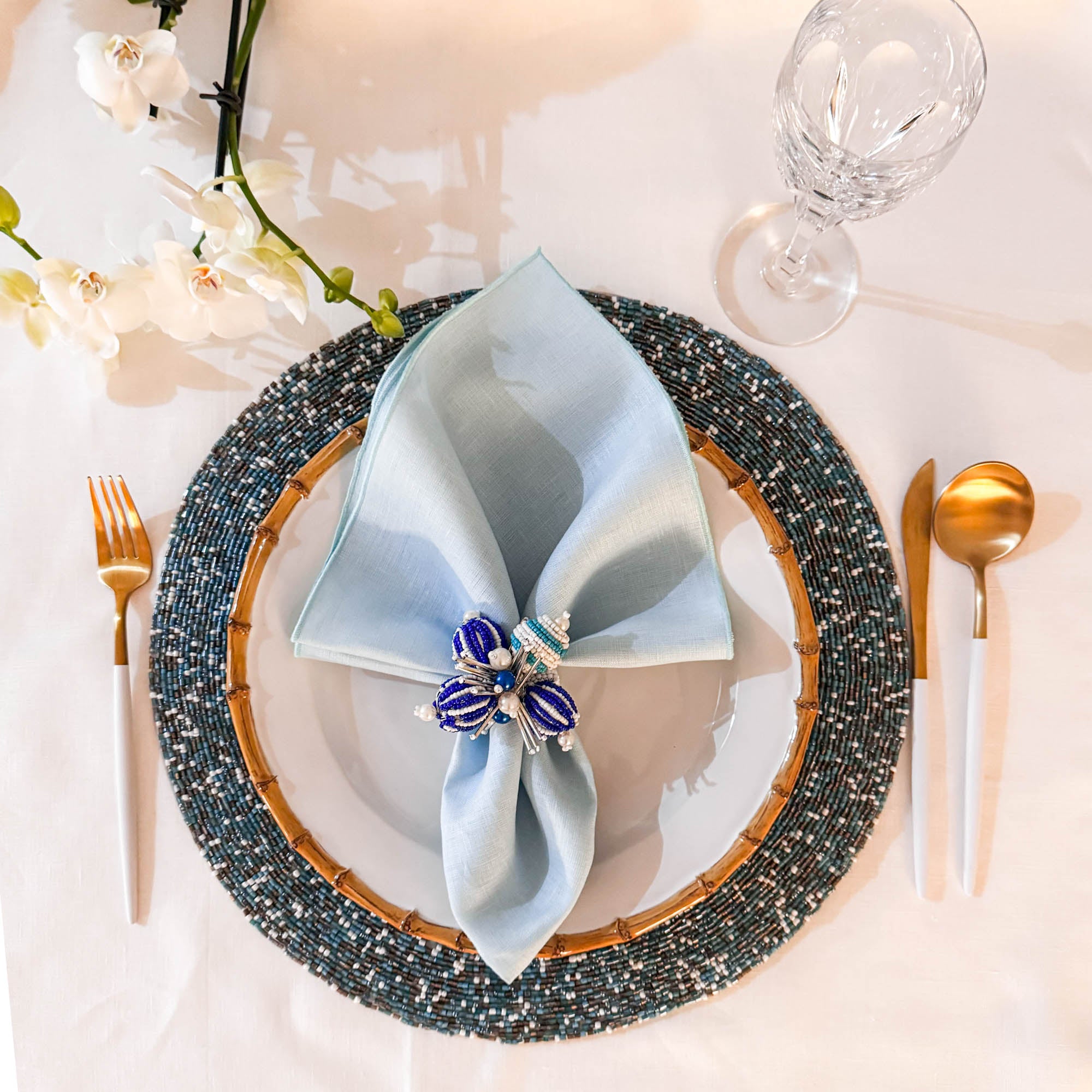 Elegant table setting with a blue linen napkin, gold cutlery, beaded napkin ring, placemat and decorative elements.