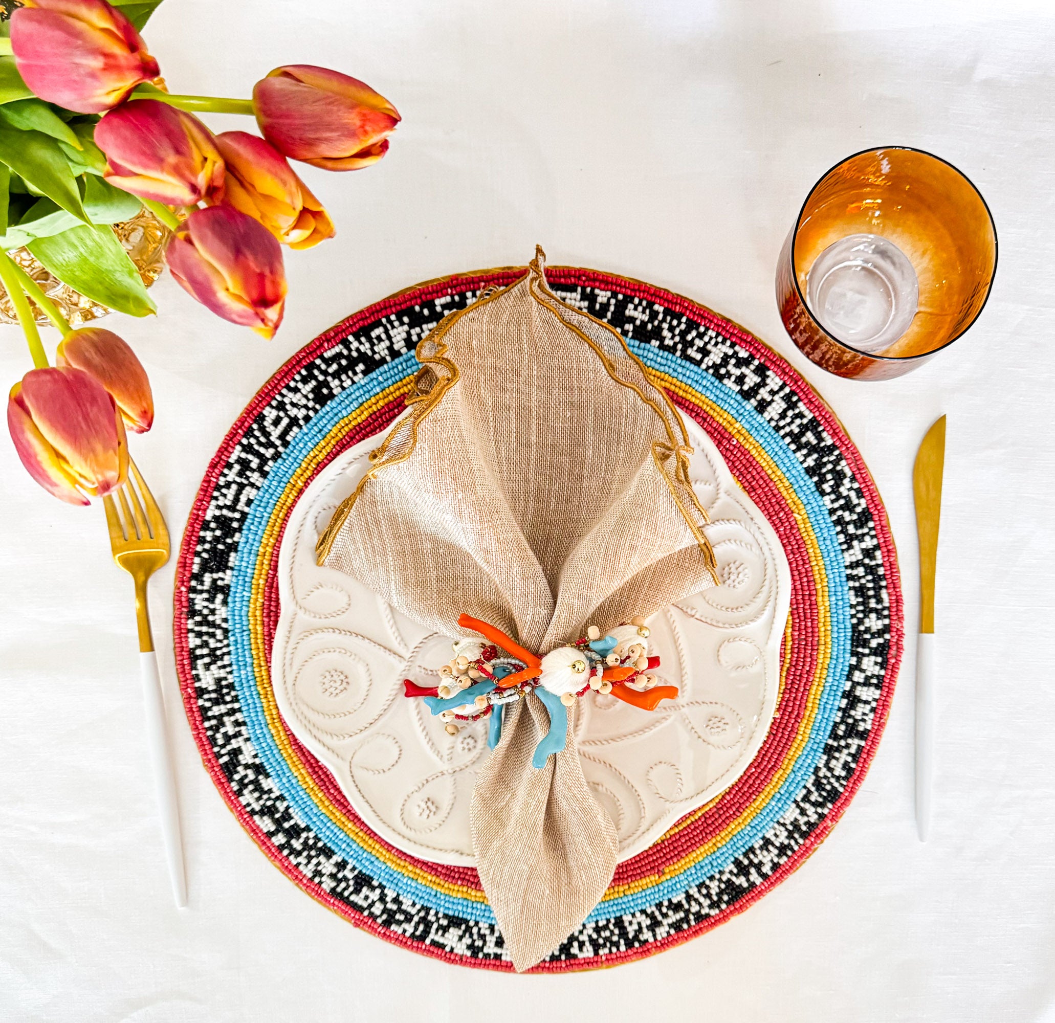 Beige linen napkin with gold ruffled edge styled on a colorful decorative plate with beaded napkin ring, tulips and glassware, elegant table setting