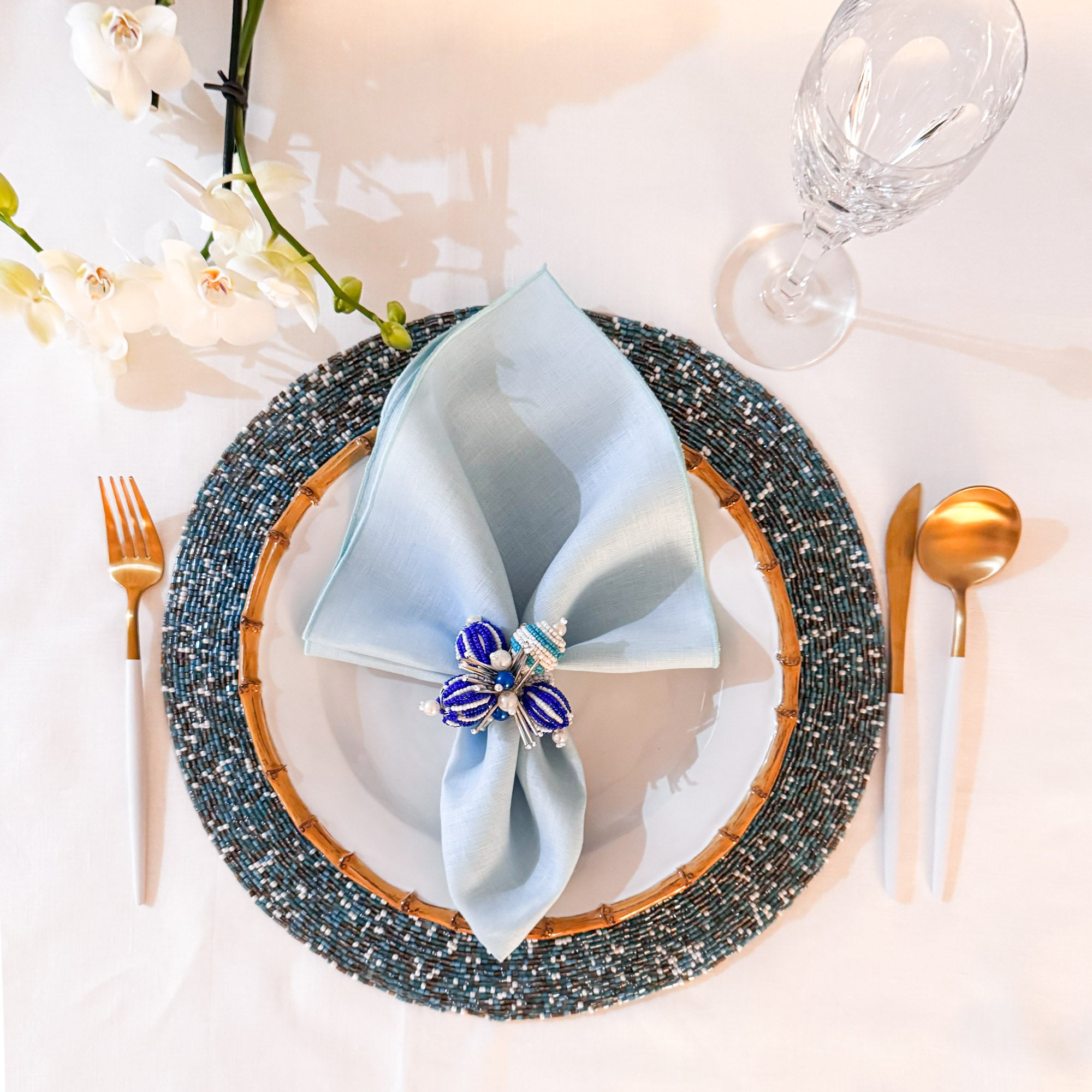 green linen napkin folded on a white plate with decorative napkin ring, placed on a round beaded multicolor placemat perfect four luxury weddings, dinners, and holiday events