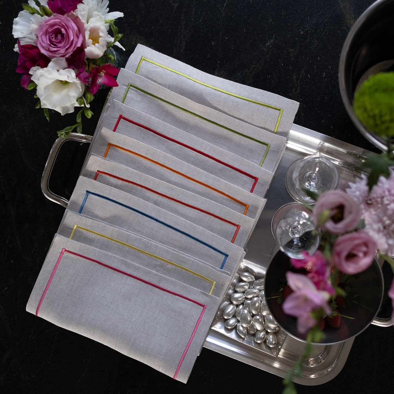 White linen cocktail napkins with colorful stitched borders displayed on a silver tray with fresh pink and white florals, elegant table styling, and vibrant embroidery edge details
