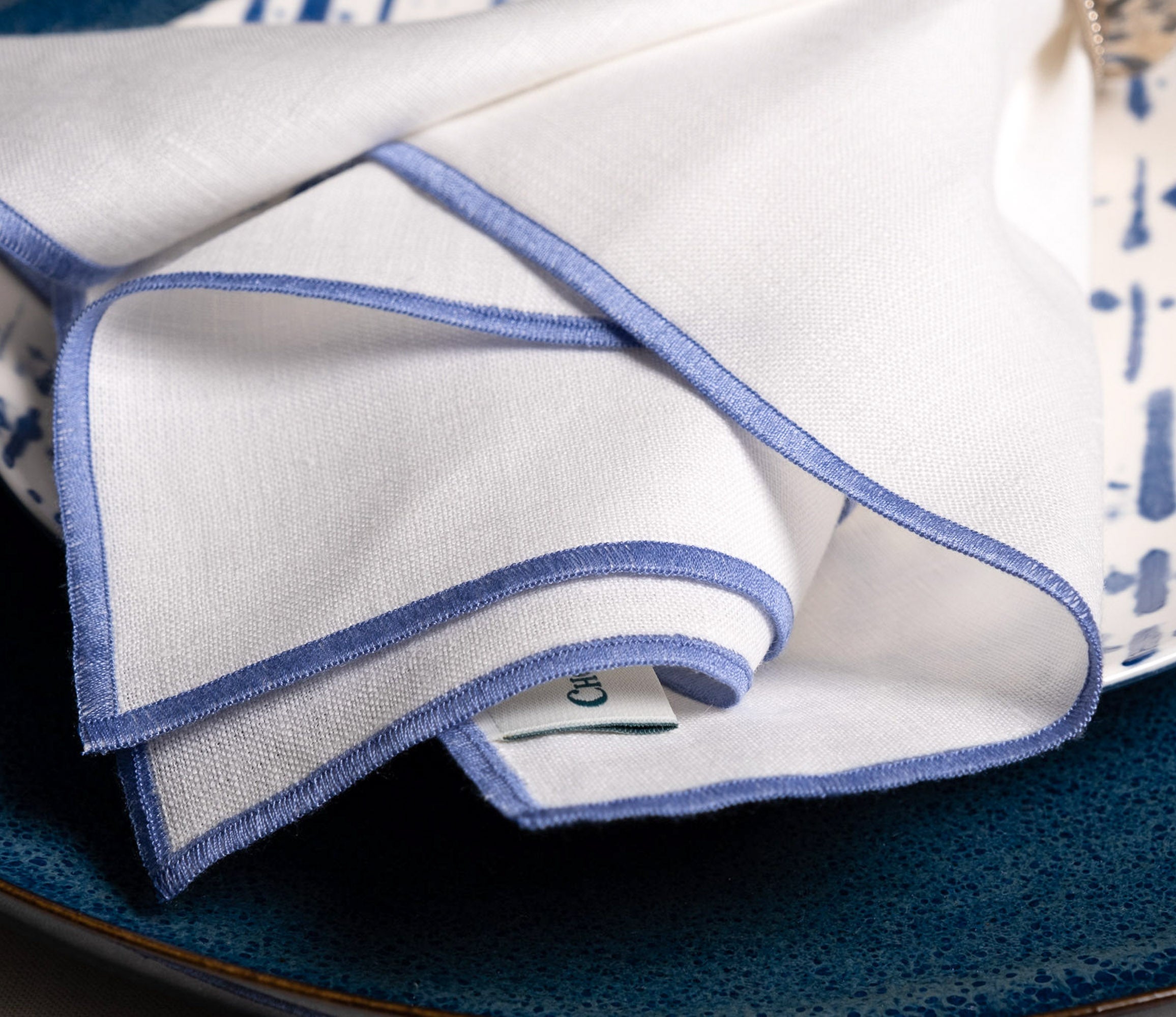 Close-up of elegant white linen napkins with delicate blue stitched edges layered over textured blue dinnerware, highlighting refined craftsmanship and timeless table styling details.