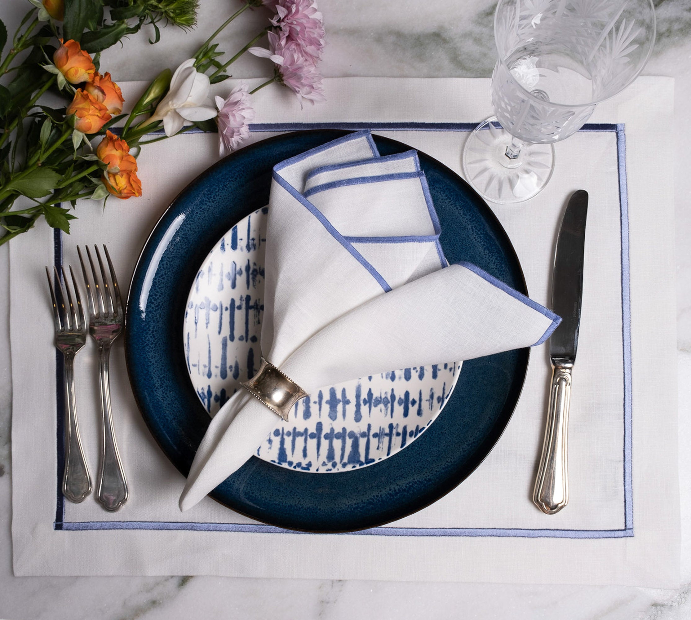 Elegant white linen table setting with blue stitched edge napkins, layered blue plates, silverware, and floral accents for a refined dining experience