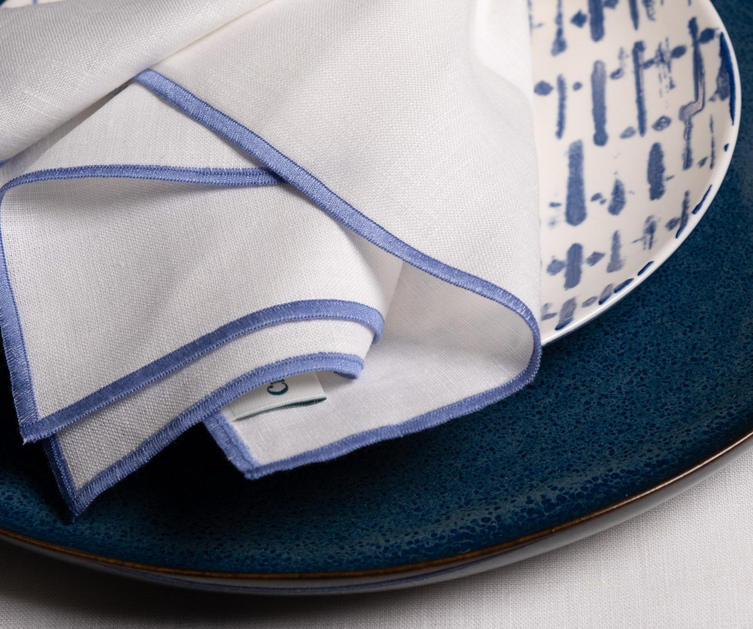 Close-up of elegant white linen napkins with delicate blue stitched edges layered over textured blue dinnerware, highlighting refined craftsmanship and timeless table styling details.