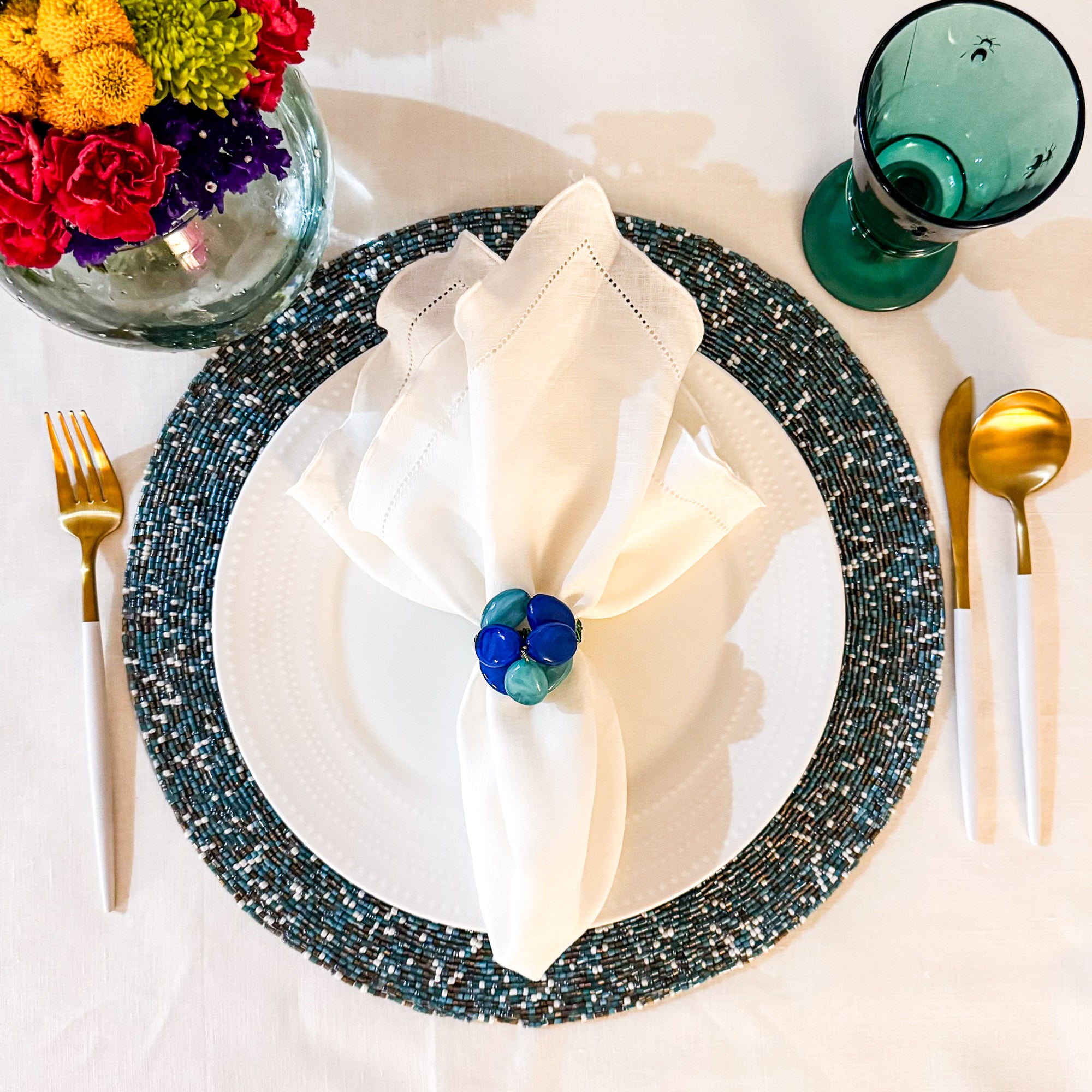 White linen napkin with ruffle hemstitched detail, layered with blue napkin ring and beaded placemat for a sophisticated dining setting.