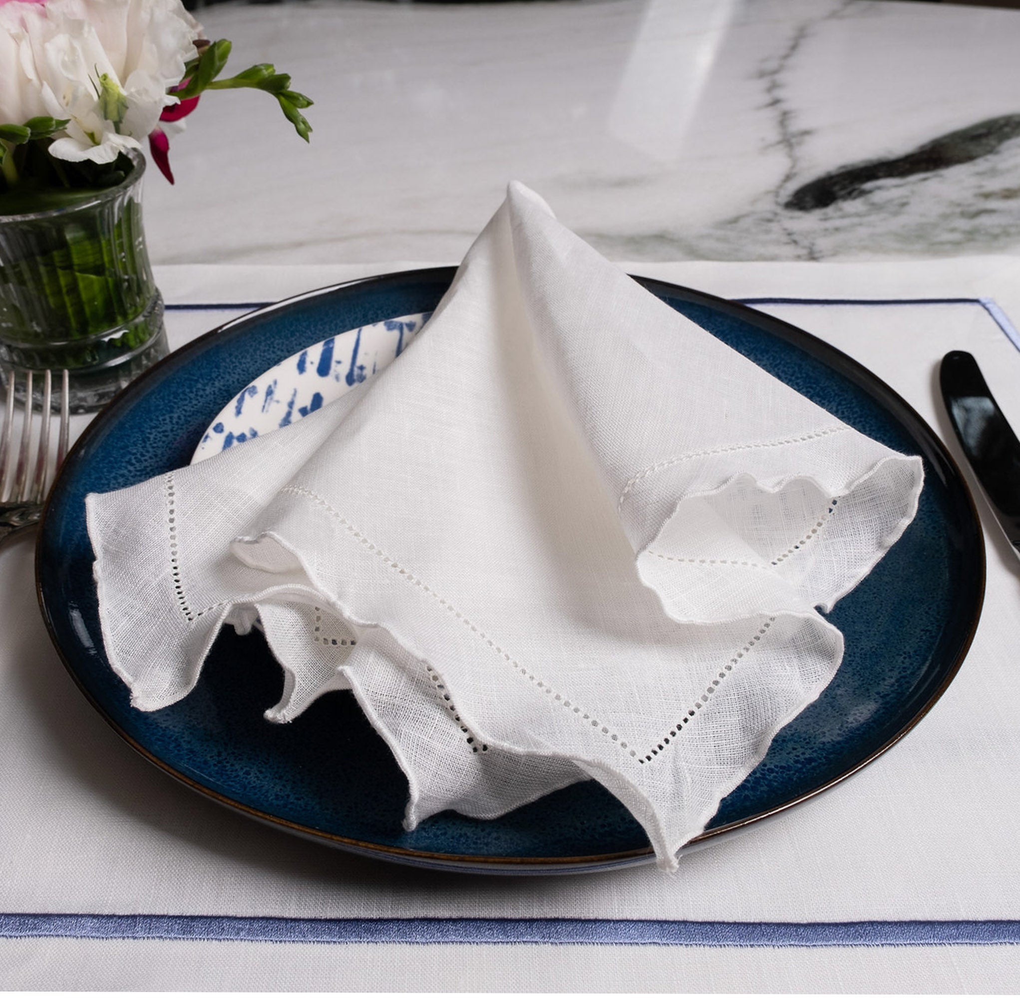 White linen napkin with ruffle hemstitched edges, elegantly folded on a blue dinner plate with matching blue bordered placemat.