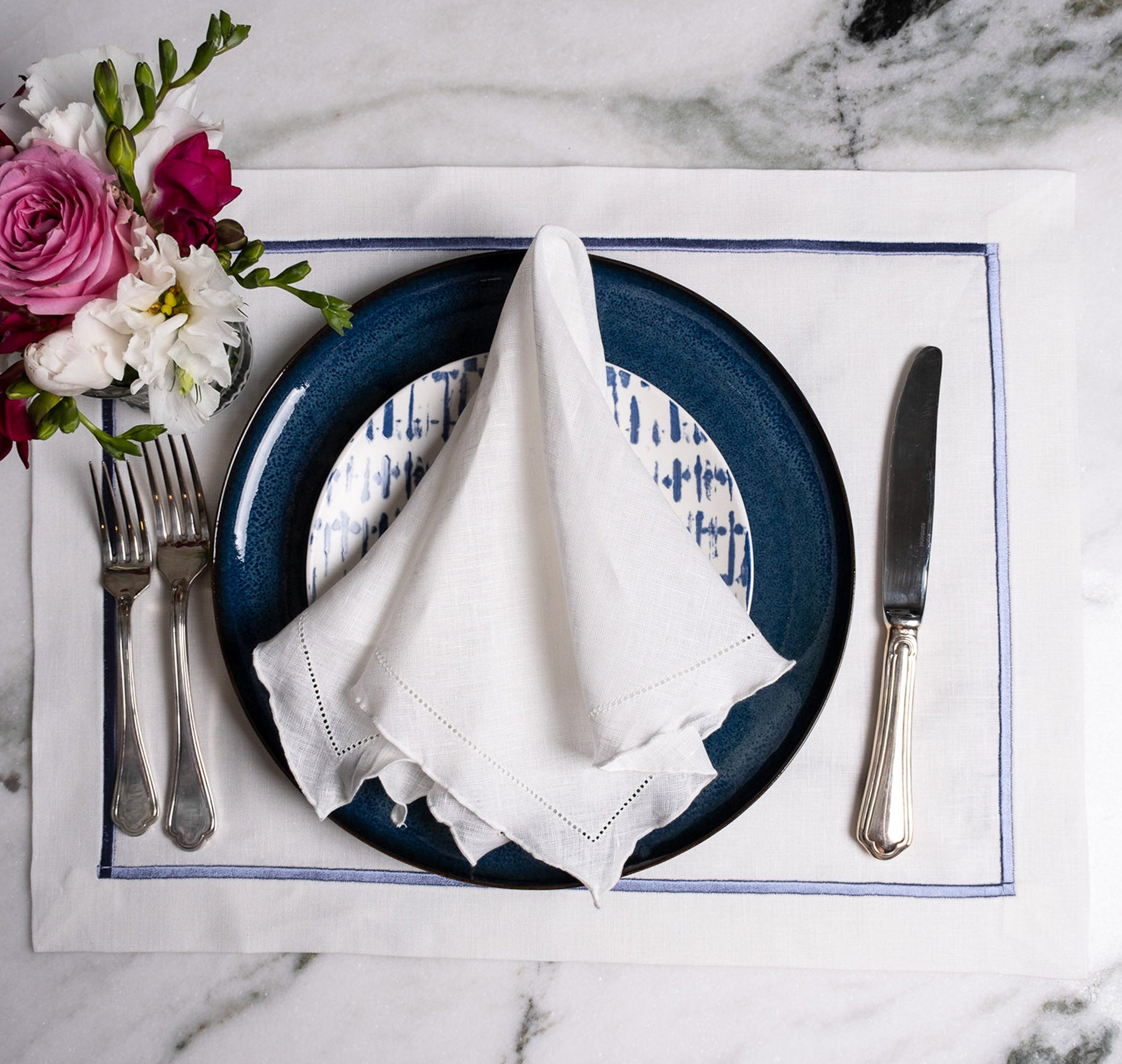 legant white linen napkin with ruffle hemstitched edges, styled on a blue patterned dinner plate with matching blue bordered placemat.